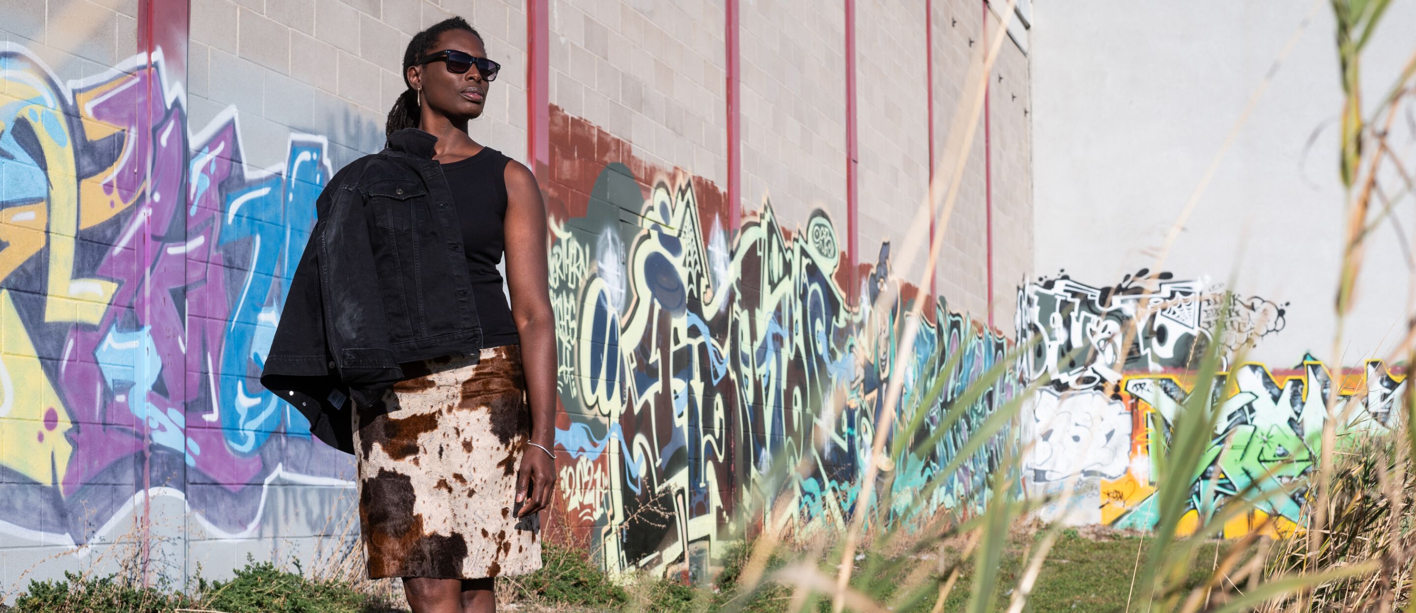 Tricia Lake, in sunglasses and a cow print skirt, exterior with graffiti wall in background.