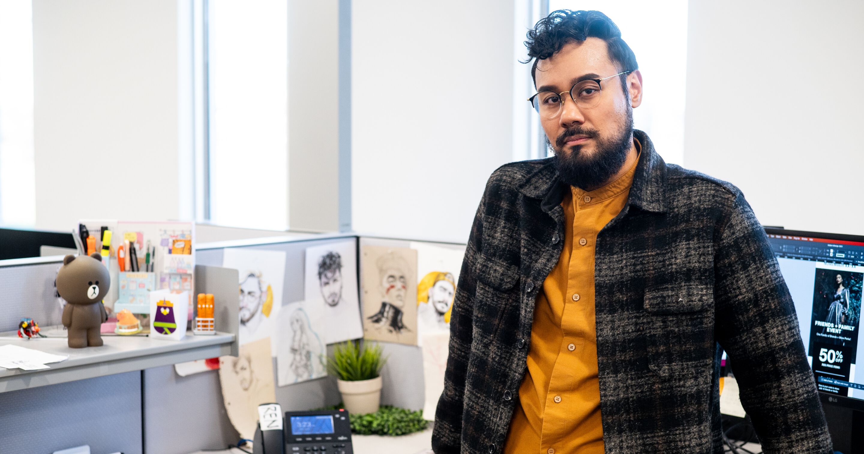 Renyl Lantano, man with a moustache, glasses, and a plaid shirt stands at desk decorated with sketches.